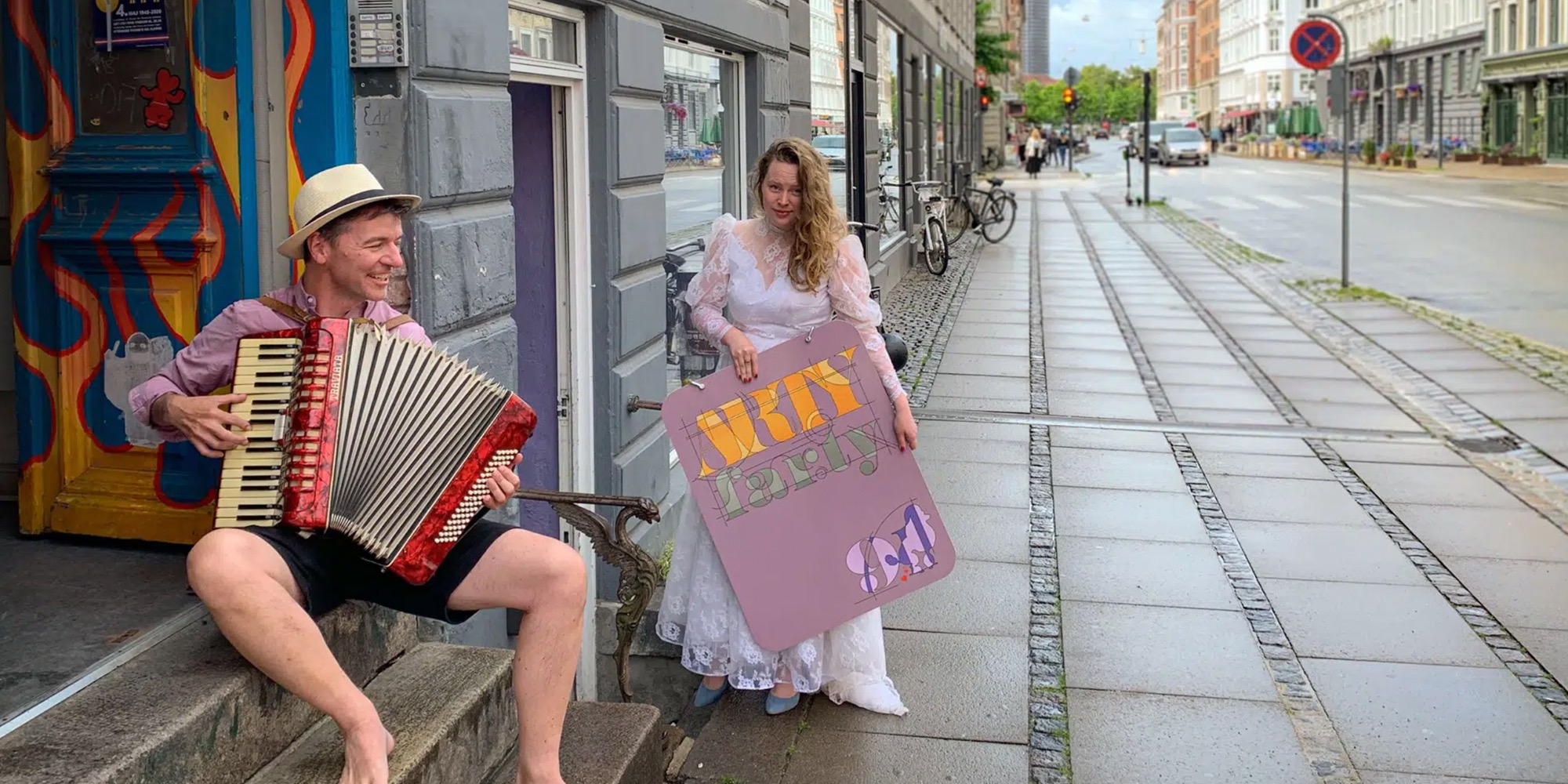 En mand med harmonika og en kvinde med skilt foran en bygning